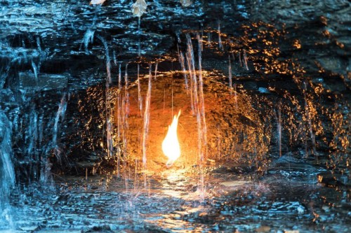 A glowing flame surrounded by water cascading over dark rocks in a cave-like setting.