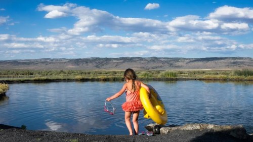 A child in a pink dress wades into a calm lake, holding sunglasses and a yellow inflatable ring, with mountains in the background.