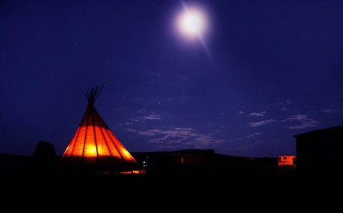 A glowing teepee under a bright moon in a starry night sky.