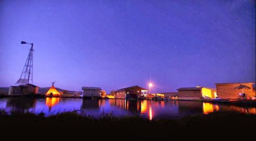 A serene night scene of wooden structures by a calm water body, illuminated under a starry sky.