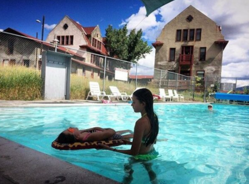 A girl in a swimsuit holds another girl on a float in a pool, with an old building and blue sky in the background.