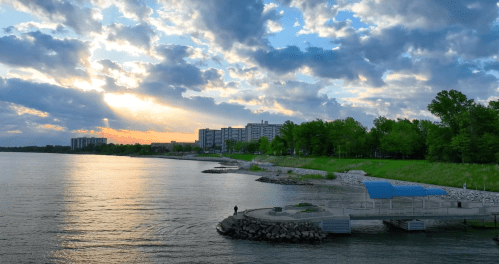 A serene waterfront scene at sunset, featuring clouds, a calm river, and a park with a walkway and buildings in the background.