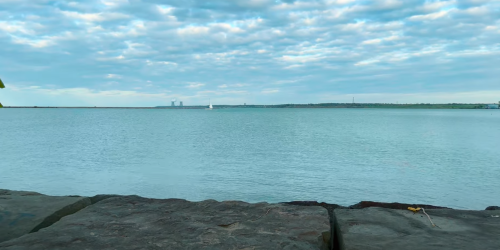 A calm lake scene with a sailboat in the distance, surrounded by clouds and a rocky shoreline.
