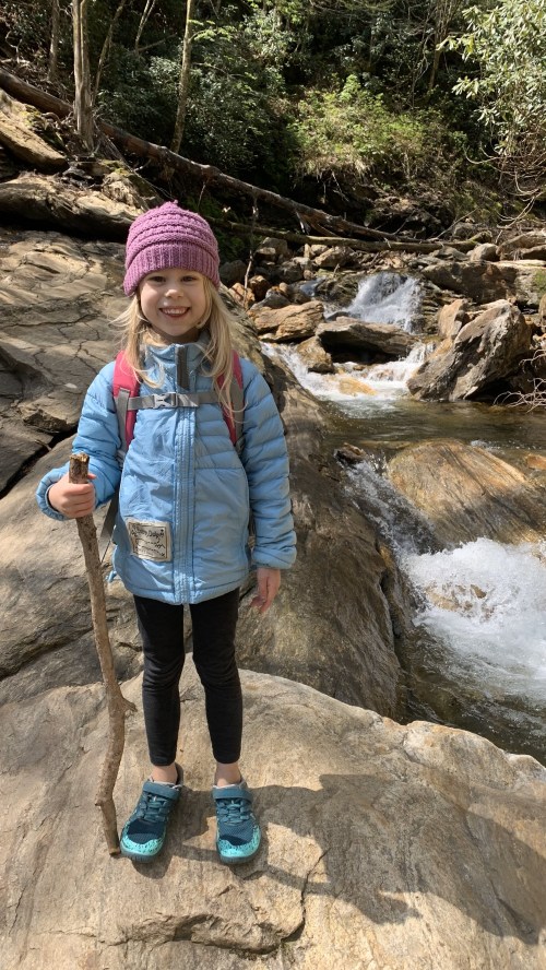 A young girl stands on a rocky riverbank, smiling and holding a stick, wearing a blue jacket and purple hat.