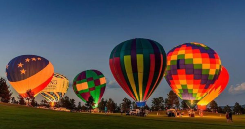 Colorful hot air balloons illuminated against a twilight sky, lined up on a grassy field.