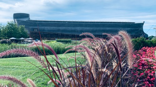 A large wooden structure resembling Noah's Ark, surrounded by greenery and colorful flowers under a cloudy sky.