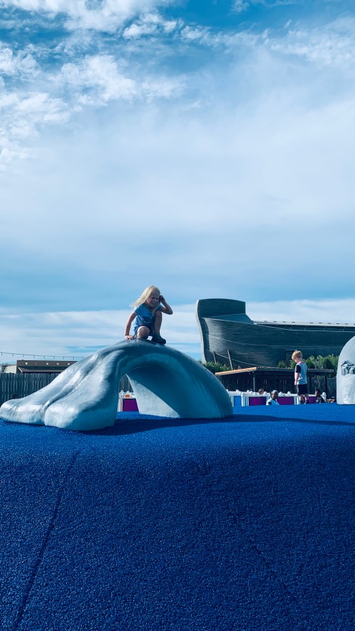 A child sits on a whale-shaped playground structure with a large building in the background under a blue sky.