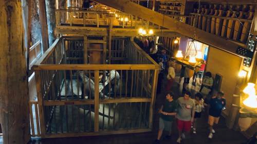 Interior view of a rustic building with wooden structures, featuring a caged area and people walking around.