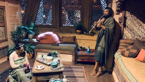 A man plays a flute while a woman crafts in a cozy, rustic room filled with plants and decorative items.