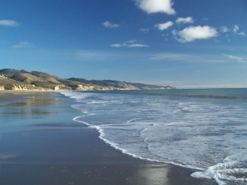 A serene beach scene with gentle waves lapping at the shore under a clear blue sky and distant hills.