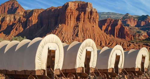 Covered wagons lined up against a backdrop of red rock formations and blue sky.