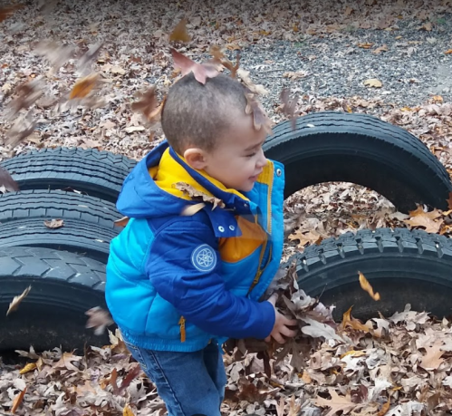 A young child in a blue jacket joyfully throws leaves in the air near a stack of tires in a fall setting.