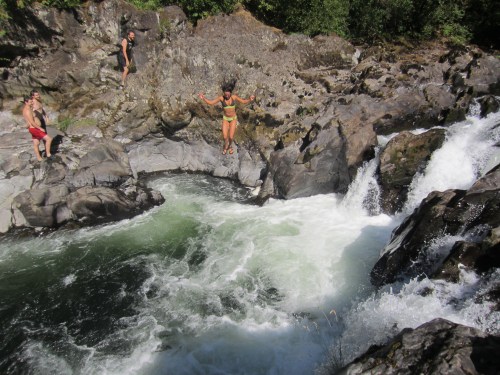 A person jumps off a rock into a river, while two others stand nearby on the rocky shore. Water cascades around them.