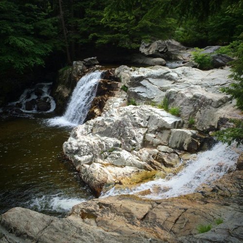 A serene waterfall cascading over rocky terrain, surrounded by lush green trees and a calm pool of water below.