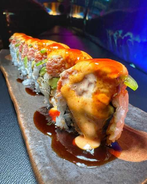 A colorful sushi roll topped with spicy sauce, served on a dark plate against a vibrant background.