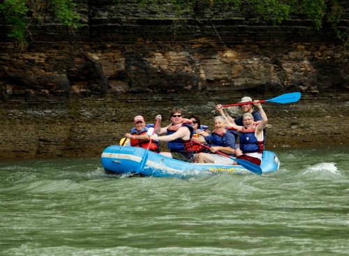 A group of people in life jackets paddling a blue raft on a river, surrounded by rocky cliffs and greenery.