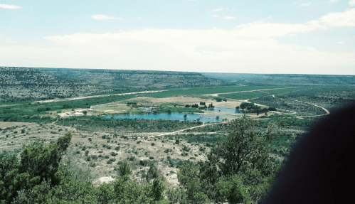 A panoramic view of a green landscape with a pond, fields, and distant hills under a blue sky.