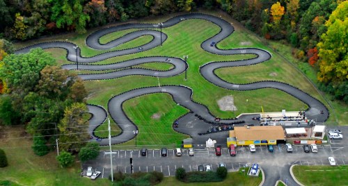 Aerial view of a winding go-kart track surrounded by trees, with karts racing and a parking lot nearby.