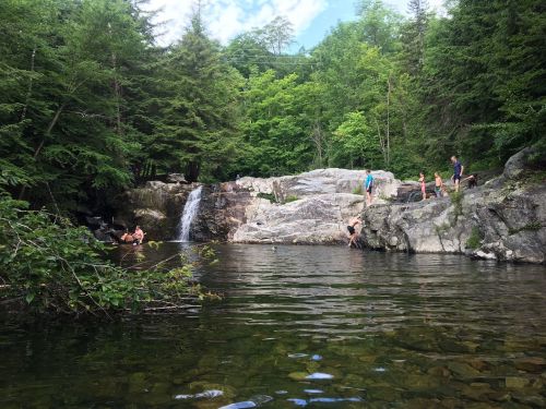 A serene natural scene with a waterfall, rocky terrain, and people swimming and enjoying the water surrounded by trees.