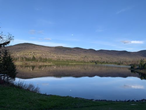 A serene lake reflecting mountains under a clear blue sky, with a hint of moon visible above the horizon.