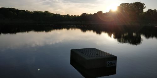 A serene lake at sunset, featuring a floating platform marked "20m" and reflections of trees and the sky on the water.