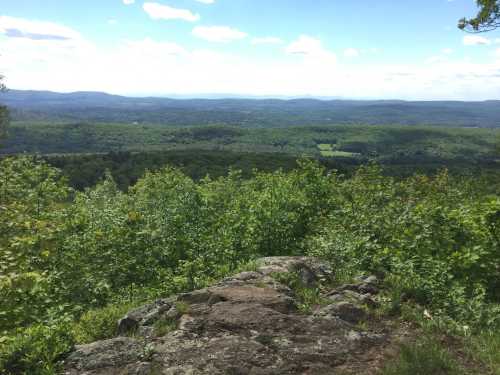 A scenic view from a rocky overlook, showcasing lush green hills and a clear blue sky with scattered clouds.