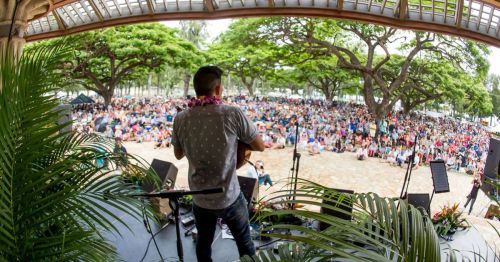 A performer on stage in front of a large crowd seated outdoors under trees.