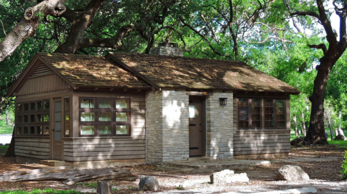 A small, rustic cabin with stone and wood exterior, surrounded by lush green trees and dappled sunlight.