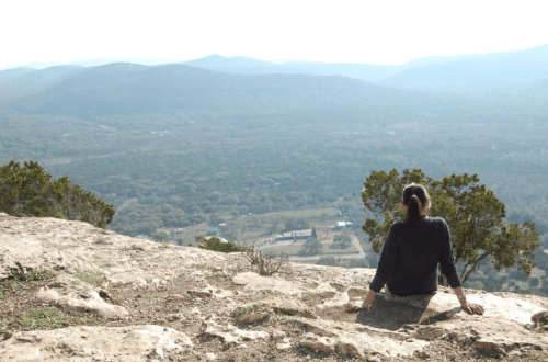 A person sits on a rocky ledge, gazing at a vast mountainous landscape under a clear sky.