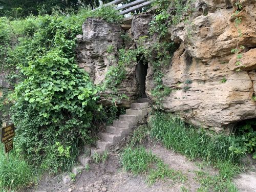 A stone staircase leads into a rocky cave, surrounded by lush green vegetation and a wooden sign nearby.