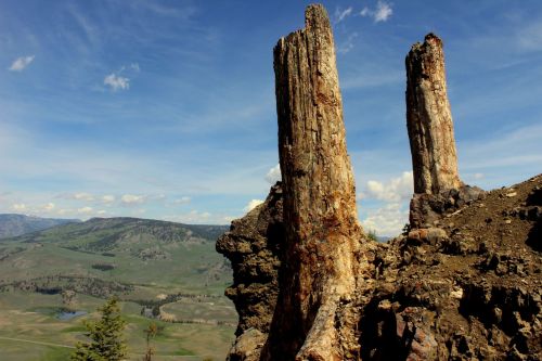Two tall, weathered rock formations stand against a blue sky, overlooking a lush green valley and distant mountains.