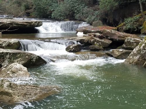 A serene river flows over rocky ledges, surrounded by lush greenery and trees. Gentle cascades create a peaceful scene.