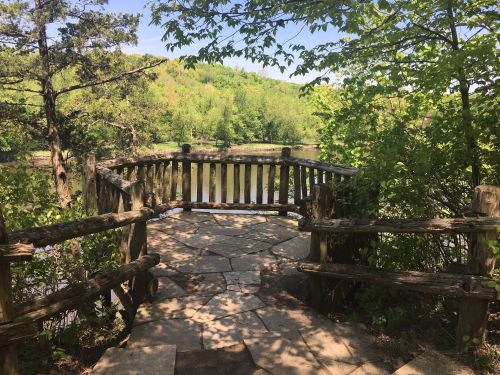A wooden overlook with a stone path, surrounded by lush greenery and a view of a calm river.