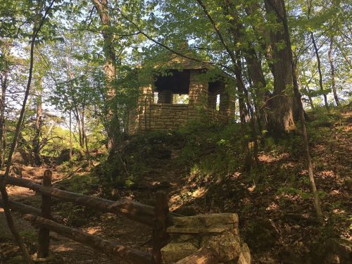A small stone structure nestled among trees on a wooded path, surrounded by greenery and sunlight.