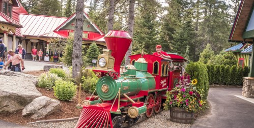 A colorful vintage train sits in a scenic area surrounded by trees and flowers, with people walking nearby.