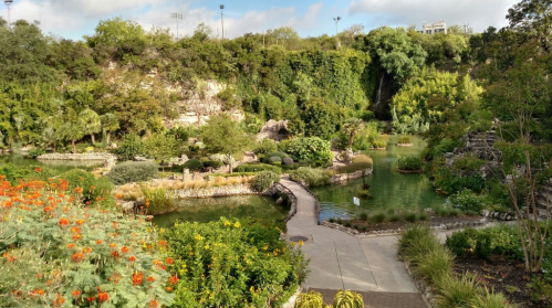 Lush garden with vibrant flowers, winding paths, and a serene pond surrounded by greenery and rocky cliffs.