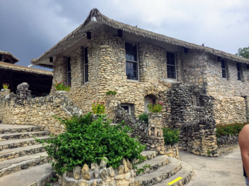 A stone building with a thatched roof, surrounded by greenery and steps, set against a cloudy sky.
