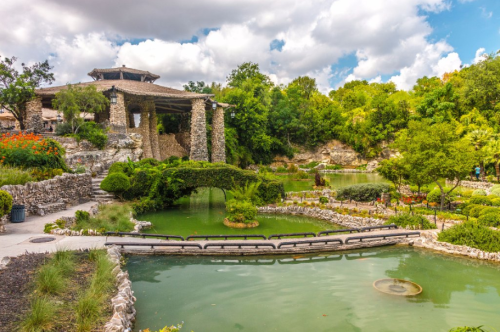 A serene garden with a pond, stone bridge, and lush greenery under a partly cloudy sky.