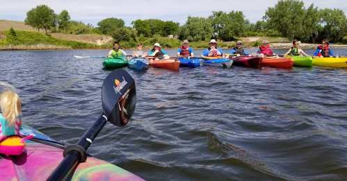 A group of people in colorful kayaks on a calm river, with trees and grassy banks in the background.