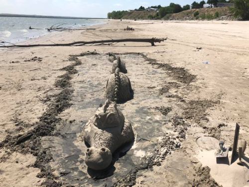 Sand sculptures resembling sea creatures on a beach, with a muddy shoreline and distant trees in the background.
