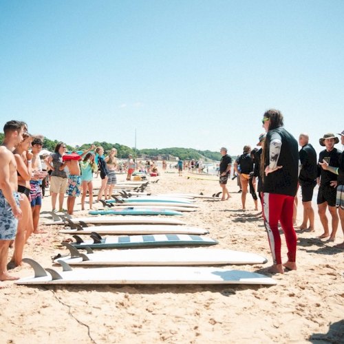 A group of people on a beach, gathered around surfboards, with a surf instructor speaking to them under a clear blue sky.
