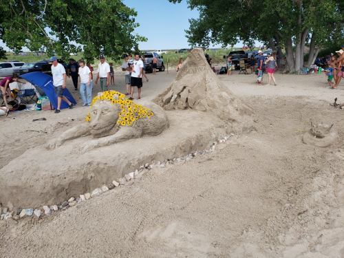 A sandy sculpture of a lioness adorned with sunflowers, surrounded by beachgoers and trees.
