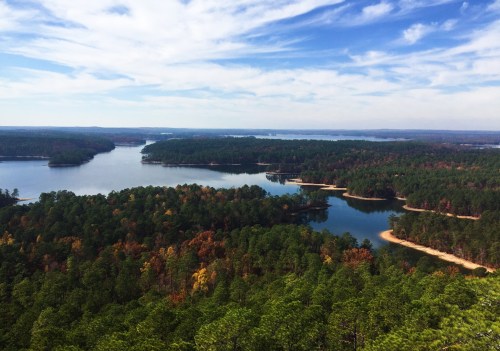 A scenic view of a lake surrounded by lush green forests and colorful autumn foliage under a blue sky.