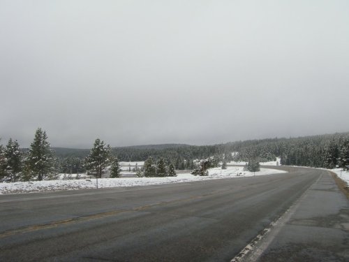 A snowy landscape along a winding road, with trees and a cloudy sky in the background.