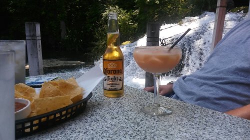 A refreshing drink and a bottle of beer sit on a table beside a plate of nachos, with a waterfall in the background.