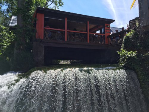 A waterfall cascades beneath a wooden structure with orange accents, surrounded by greenery and blue sky.