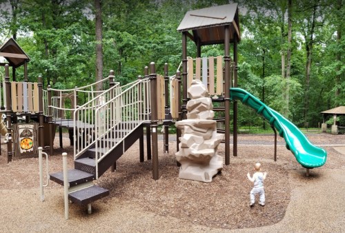 A playground with a slide, climbing structure, and a child exploring the area surrounded by trees.