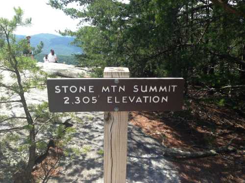 Sign at Stone Mountain Summit indicating an elevation of 2,305 feet, surrounded by trees and mountains in the background.