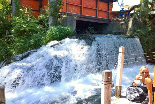 A waterfall cascades over rocks near a wooden structure, surrounded by lush greenery and a sunny atmosphere.