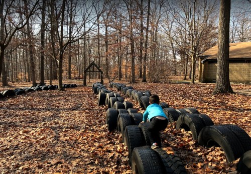 A child climbs on a path made of tires in a wooded area with fallen leaves and a wooden structure in the background.
